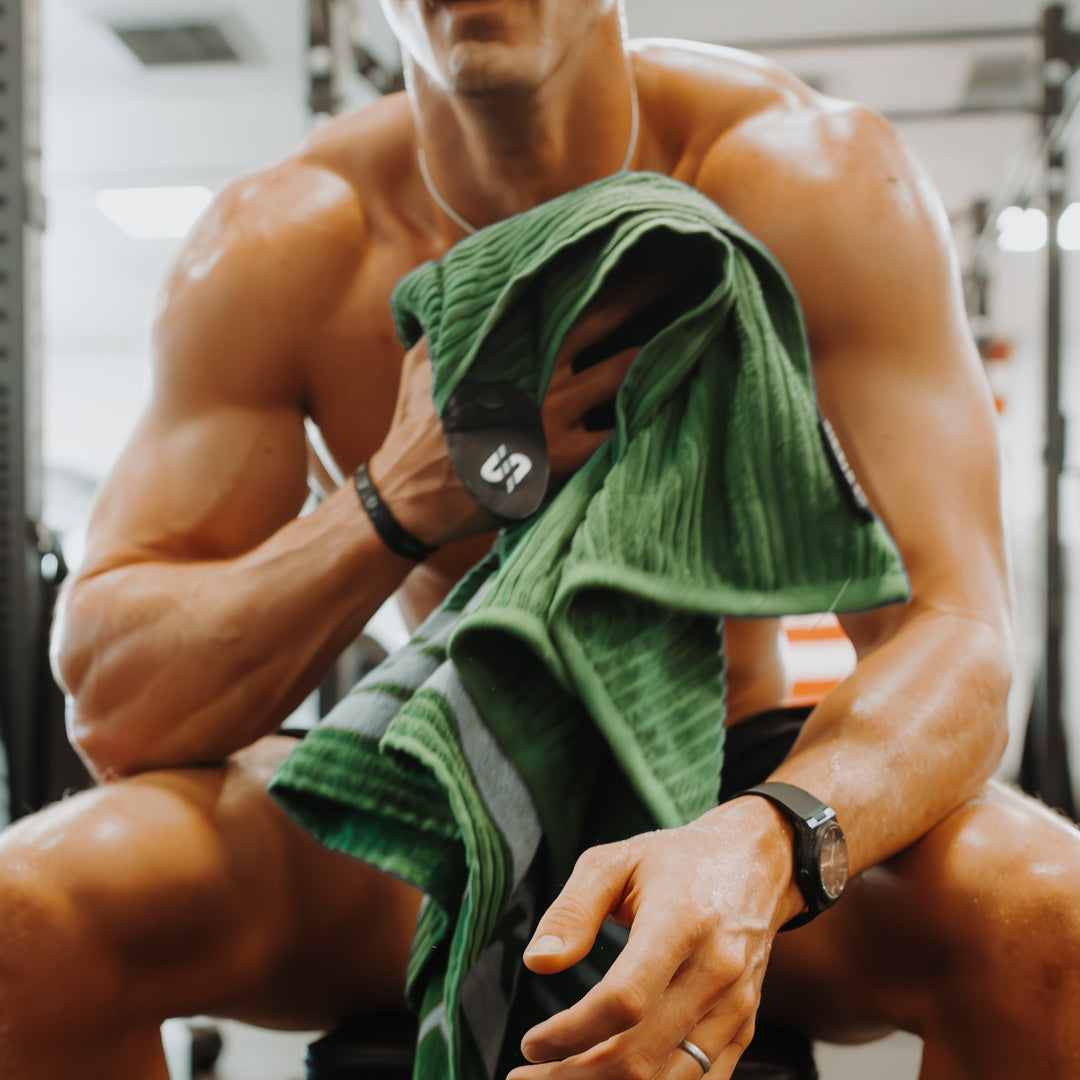 Muscular person at a gym sitting on a Rogue weight bench with a STICKIT green XL towel