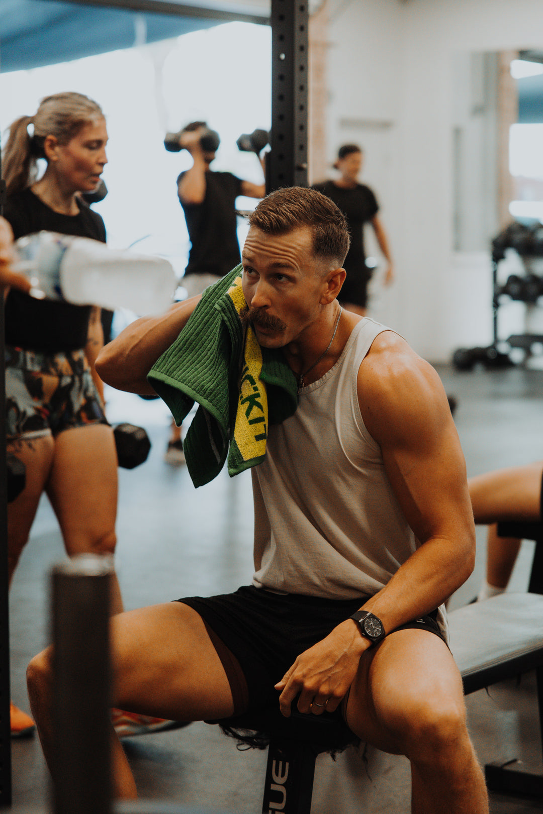 Muscular man in a gray tank top and black shorts, wiping sweat with a green towel in a modern gym setting.