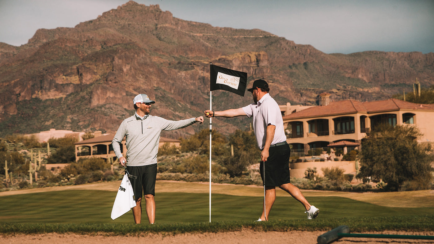 Two male golfers in light gray and white attire enjoying a sunny day on a green golf course with mountains in the background.