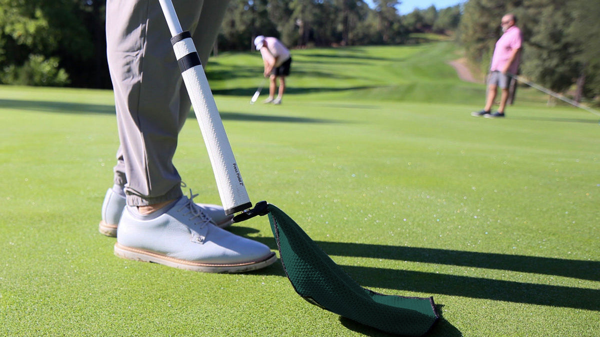 Men's golf shoes in light blue with beige accents, paired with a green golf towel on the putting green.