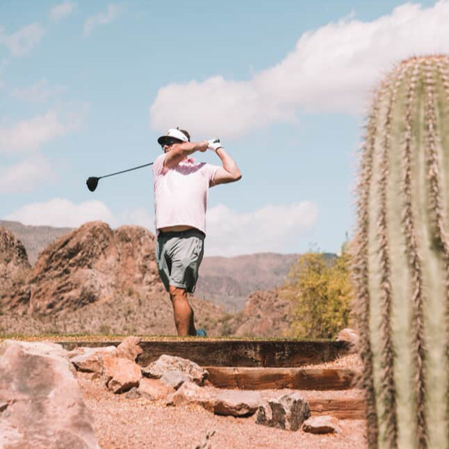 Man in a pink golf shirt and gray shorts swinging a driver club on a rocky course with cacti in the background.