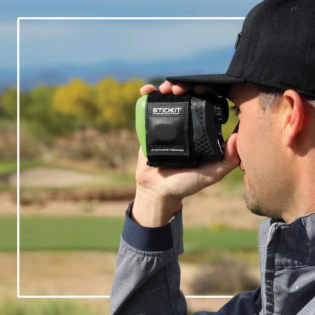 Man using a green and black StickIt golf rangefinder on a sunny day, set against a blurred outdoor landscape.