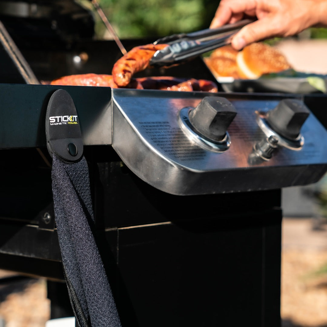 Black and stainless steel grill with a "Stick It" hanging towel holder, grilling sausages and burgers outdoors.