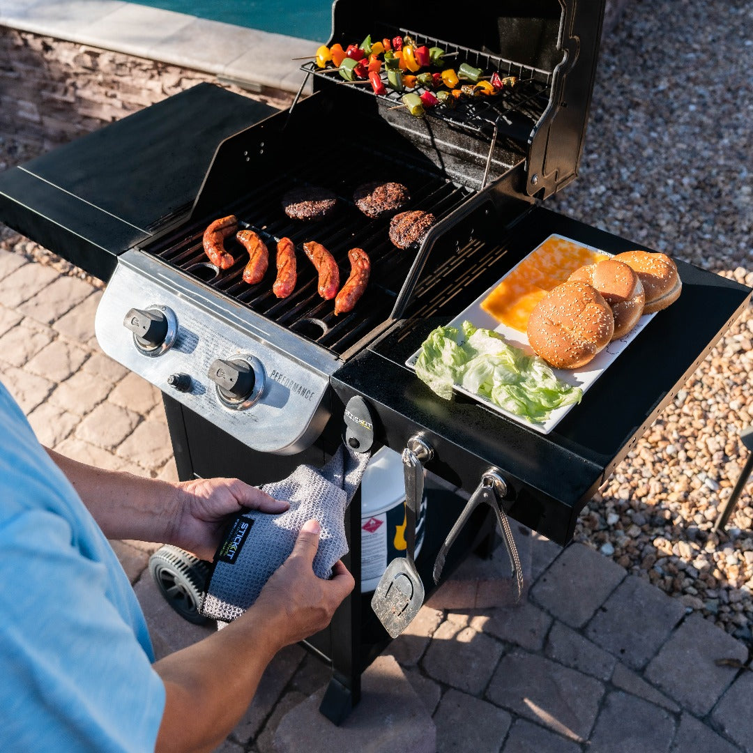 Black gas grill with sausages, burgers, and colorful vegetables cooking; outdoor patio setting, casual summer BBQ style.