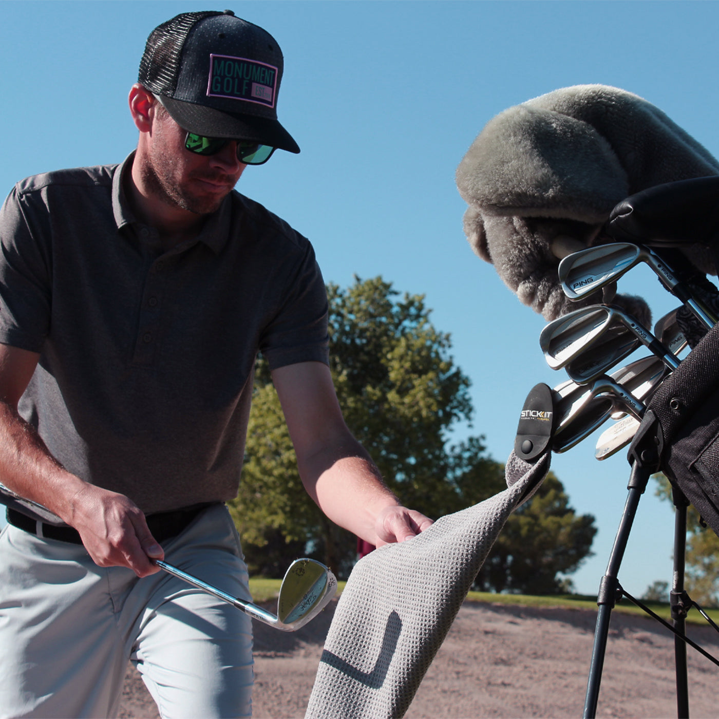 Man in gray shirt and cap sets down a gray golf towel by golf clubs on a black stand in a sunny outdoor setting.