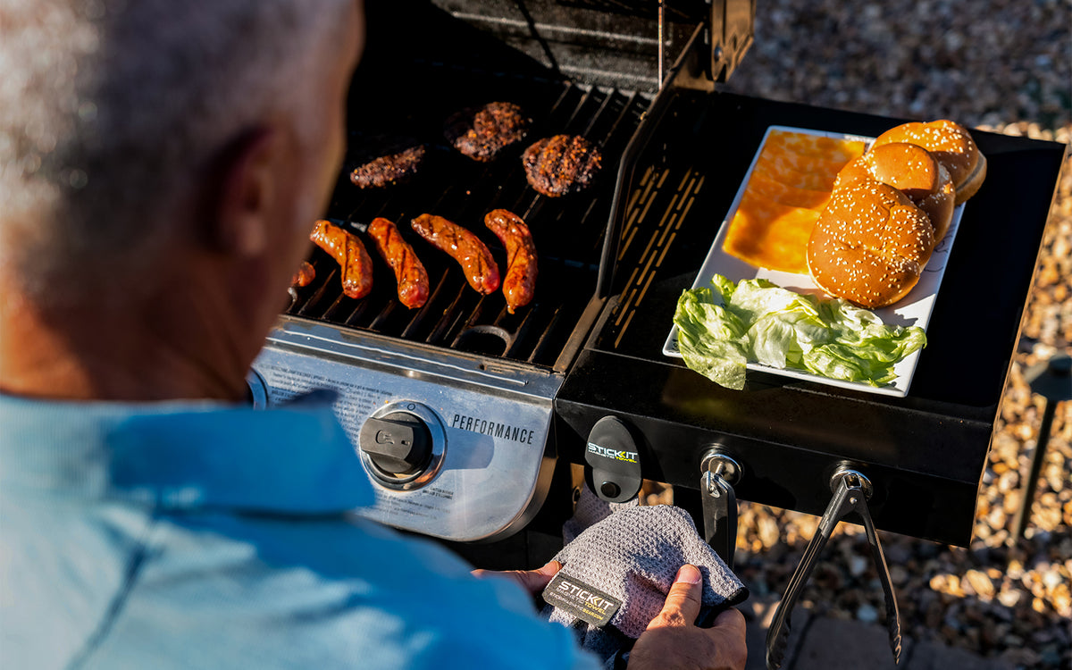 Gray grilling gloves in use, grilling burgers and sausages on a stainless steel barbecue, with fresh buns and lettuce.