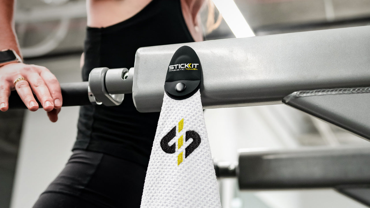 White microfiber gym towel with a black and yellow Stickit logo, hanging on a metallic gym bar.