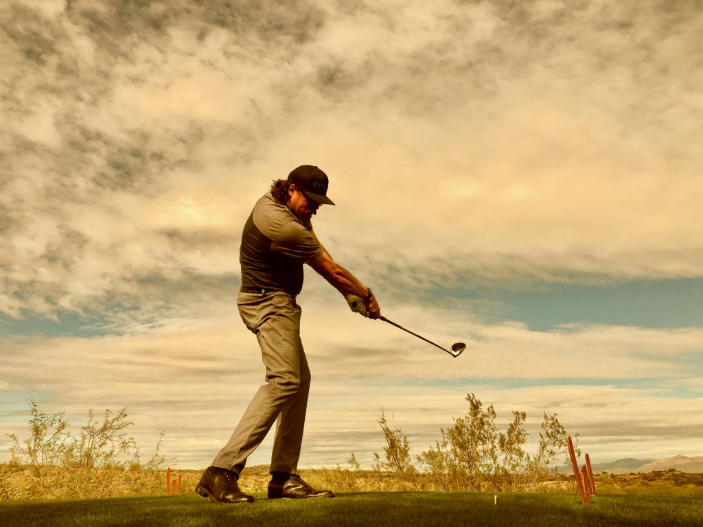 Golfer in a gray polo and light gray pants, swinging a club on a scenic course under a cloudy sky.