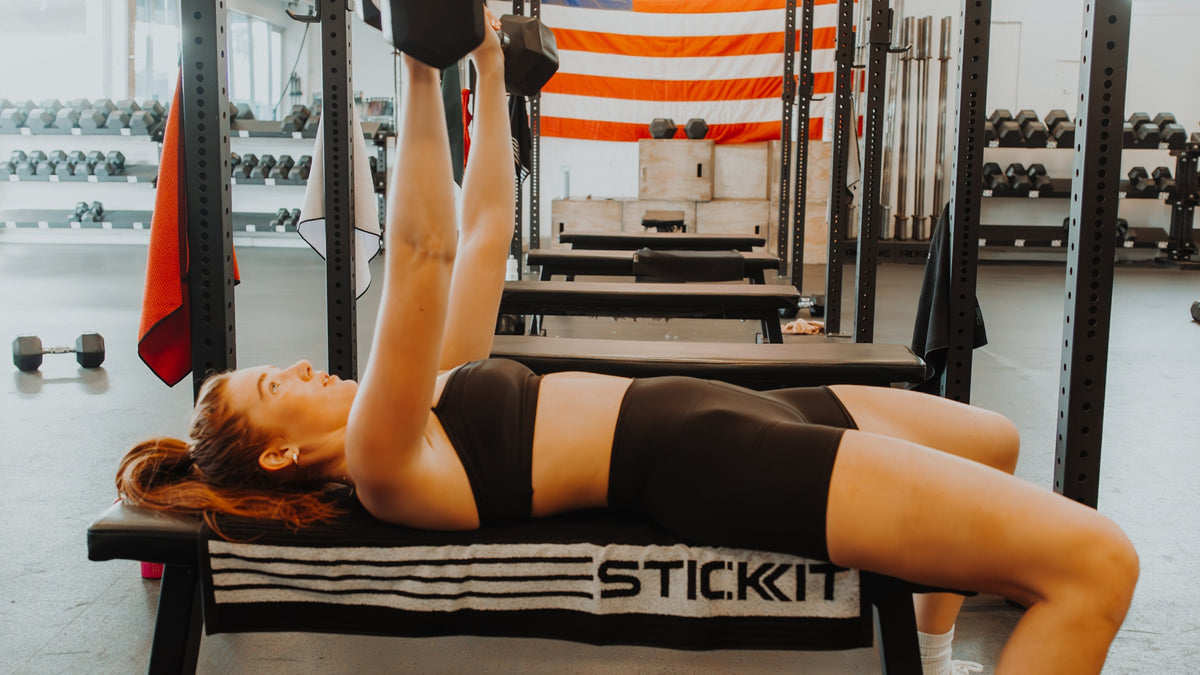 Woman exercising with dumbbells on a bench with a STICKIT towel in a gym setting