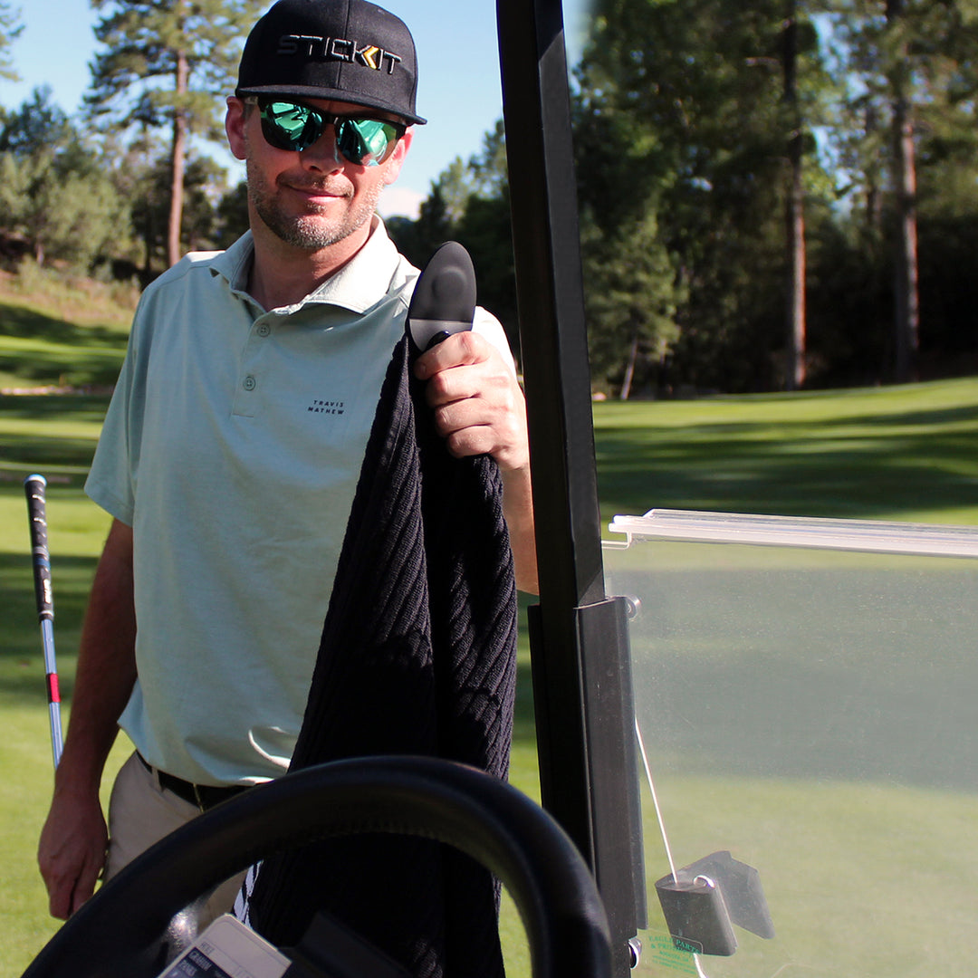 Men's light blue golf polo shirt with stylish black cap, holding a black golf towel on a sunny course.