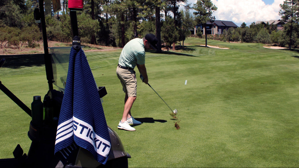 Man in a light green polo and khaki shorts swinging a golf club on a lush course, with a blue golf towel in the foreground.