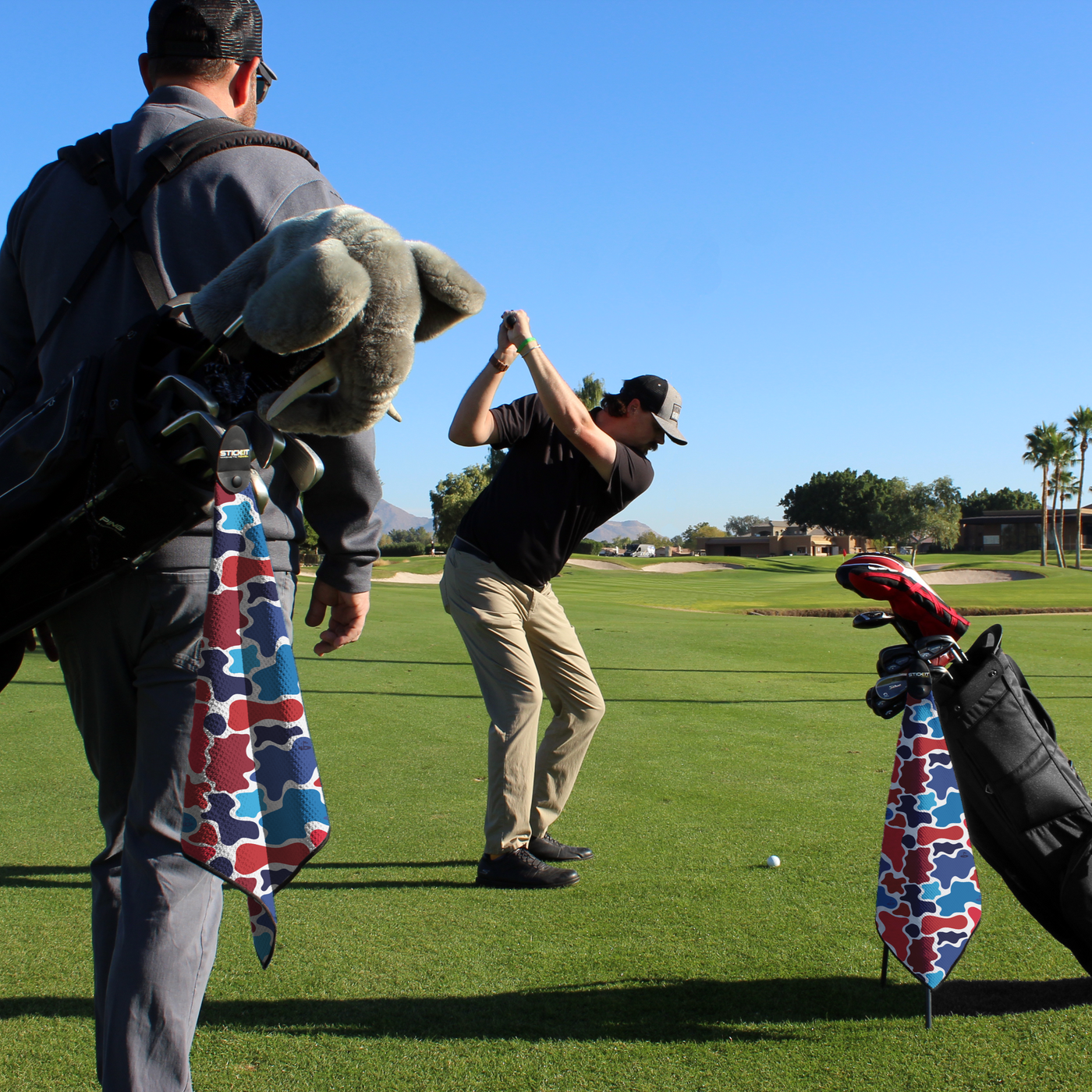 Golfer in black polo swings on green course; colorful patterned golf towels hang from clubs. Clear blue sky above.