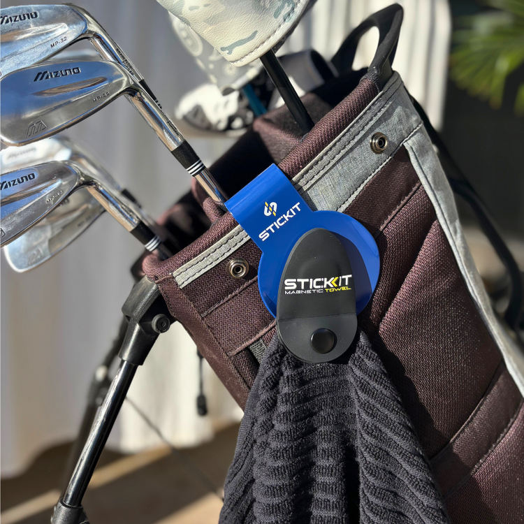 Black golf towel with a blue STICKIT clip attached to a brown golf bag, alongside stainless steel golf clubs.