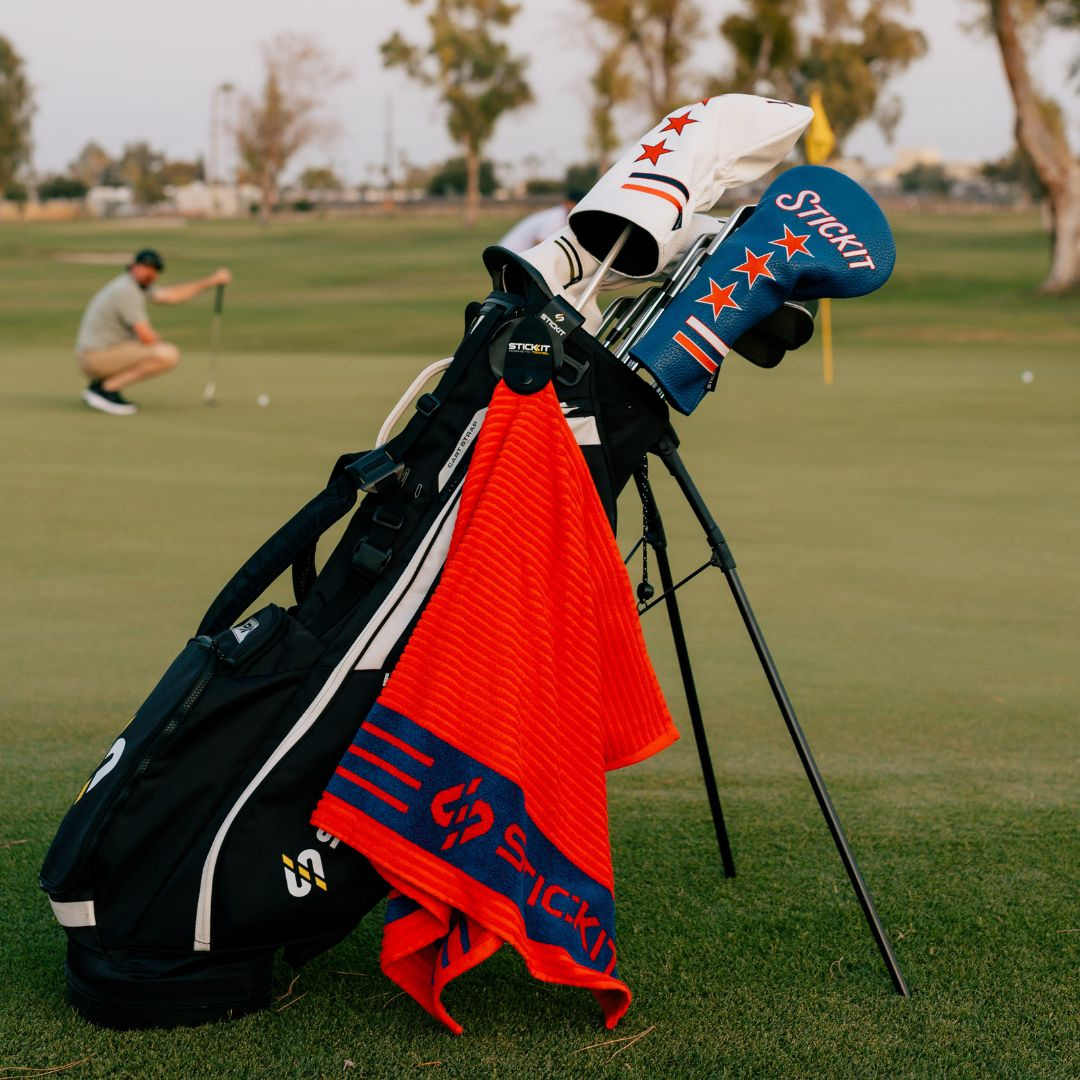 Stylish black golf bag with red and blue Sticki accessories, including headcovers and a towel, set on a lush green course.