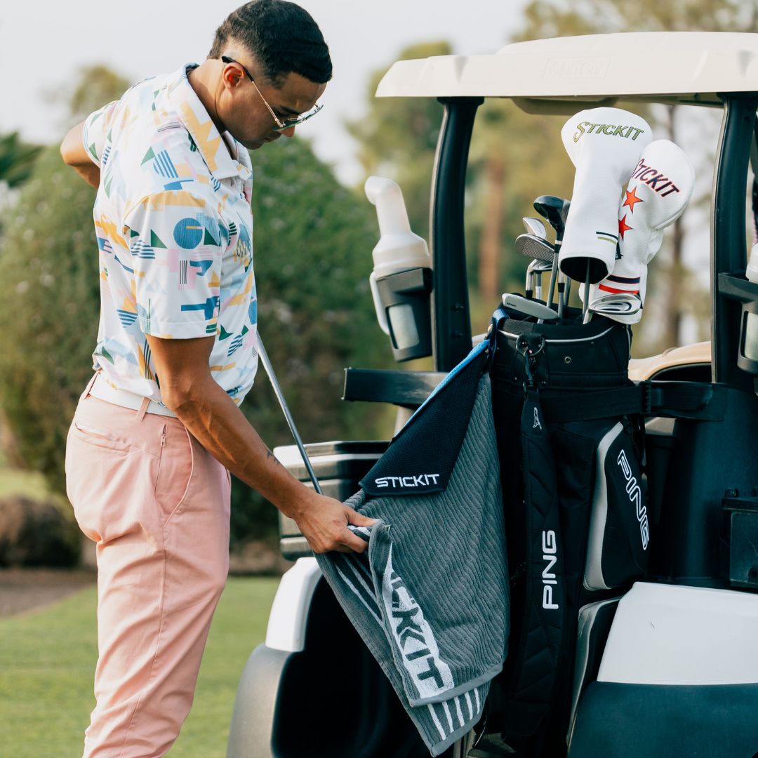 Stylish golfer in pastel shirt and pink pants retrieving gray towel from a golf cart, perfect for sporty outdoor styles.